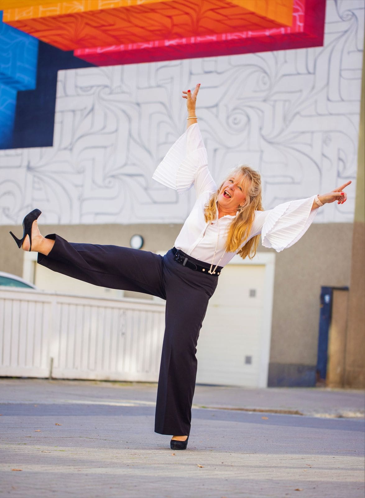 En blond kvinna skrattar och står med ett ben och båda armarna upp i luften i en glad pose.